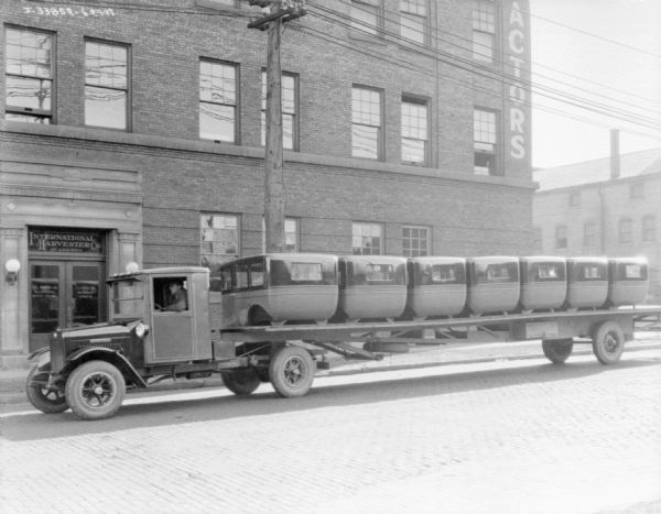 View across street towards a truck with a long truck bed loaded with bus cabs, in front of an International Harvester Co. office building.