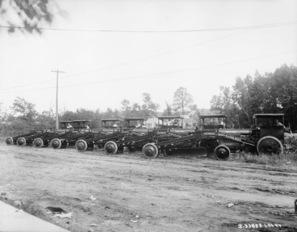View across road towards seven men, each posing in the cab of seven graders, parked at an angle in a row along the side of the road. The signs painted on the side of the graders reads: "Gilbert, Stillwater, Minnesota." Houses are in the background.