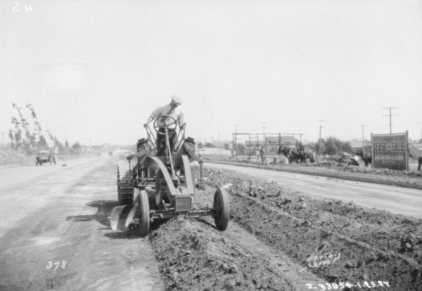 View down road towards the front of a road grader that a man is driving to level a road. A billboard on the other side of the road on the right reads, in part: "Lease or Sale, Jacobsen-Alcorn."