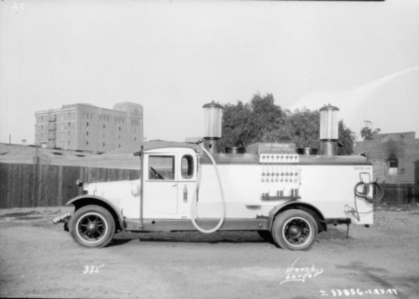 Truck parked in a yard with a fence, and buildings in the background. A sign on the back reads: "No Smoking, Stop Your Motor."
