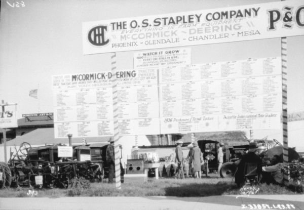 Outdoor display with a large sign for International Harvester. The large sign at the top reads: "The O.S. Stapley Company, Everything in Farm Equipment, McCormick-Deering, Phoenix, Glendale, Chandler, Mesa." Inside the display people are standing among IH trucks and tractors.