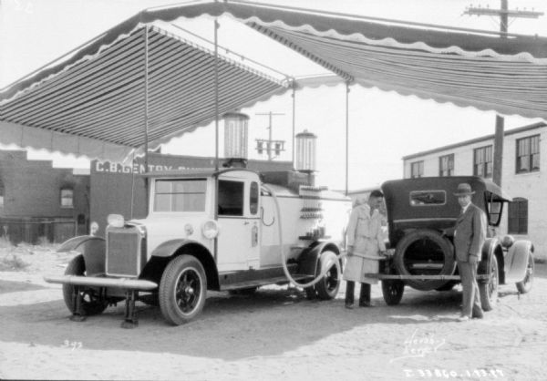 Two men are standing under a large, striped awning with a truck and an automobile. The man in the center is wearing a white coat, and is filling up the automobile from the tank of the truck with a hose. The other man is wearing a suit and hat and is standing nearby at the back of the automobile. A sign on a small building behind them reads, in part: "C.B. Gentry ___."