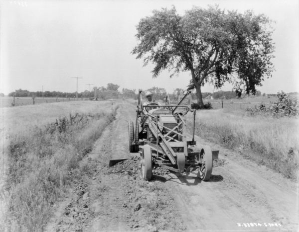 View from front of a man operating a McCormick-Deering grader on a road. There is a fence and field on the left, and on the right is a trees, and farm buildings in the distance across a field.