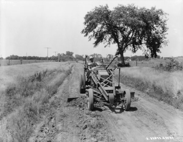 View from front of a man operating a McCormick-Deering grader on a road. There is a fence and field on the left, and on the right are trees. There are farm buildings in the distance across a field.