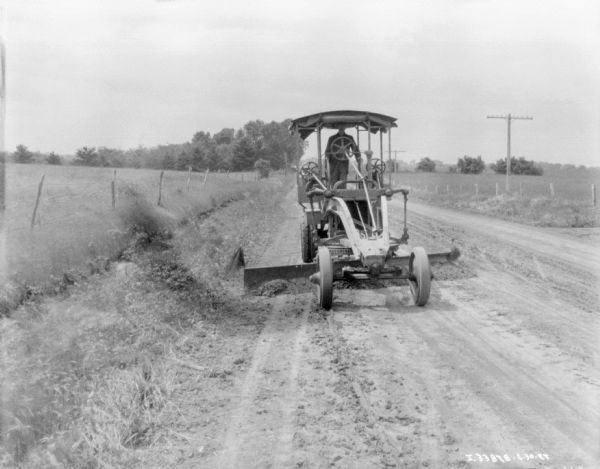 View from front of a man operating a grader on a road. There is a fence and field on the left, and more fields on the right.
