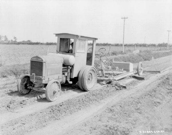 Three-quarter view from front left of a man operating a grader on a road. There is a fence and field in the background.