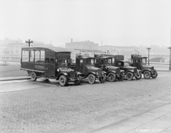 View of a fleet of delivery trucks lined up in a row. There is a bridge and buildings in the background. Signs painted on the trucks read: "Davidsons, Iowas Largest Home Furnishings."