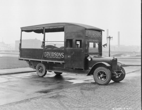 Three-quarter view from front right of a man sitting in a delivery truck. Painted on the side of the truck is a sign that reads: "Davidsons, Iowas Largest Home Furnishings." There is a bridge and buildings in the background.