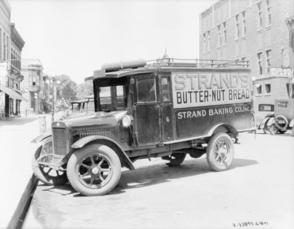 View along curb towards a bakery delivery truck parked at an angle at the sidewalk. The sign painted on the side of the trucks reads: "Strands Butter-Nut Bread, Strand Co., Inc." A spare tire is attached to the roof of the cab. Storefronts are along both sides of the street.