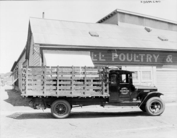 Right side view of a truck with a stake body parked in front of a building. A man is sitting in the driver's seat, and the sign painted on the passenger door reads: "Grinnell Poultry & Egg Company." The sign painted on the building reads, in part: "__LL Poultry & _."