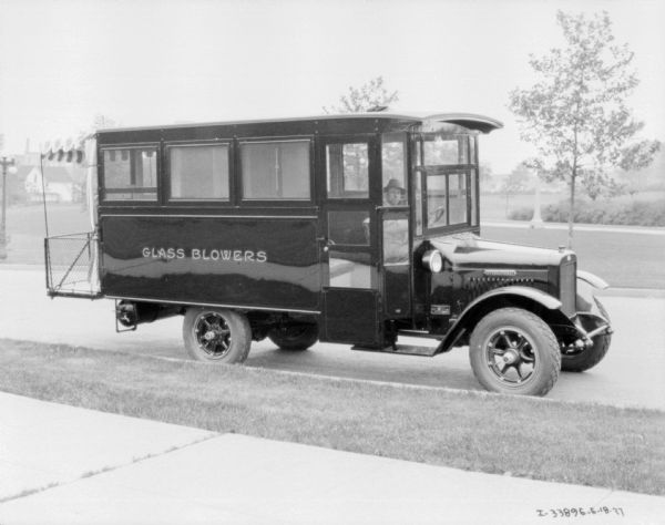 View from sidewalk towards a man sitting in the driver's seat of a truck with enclosed sides, and a platform on the back with a striped awning. The sign painted on the truck reads: "Glass Blowers."