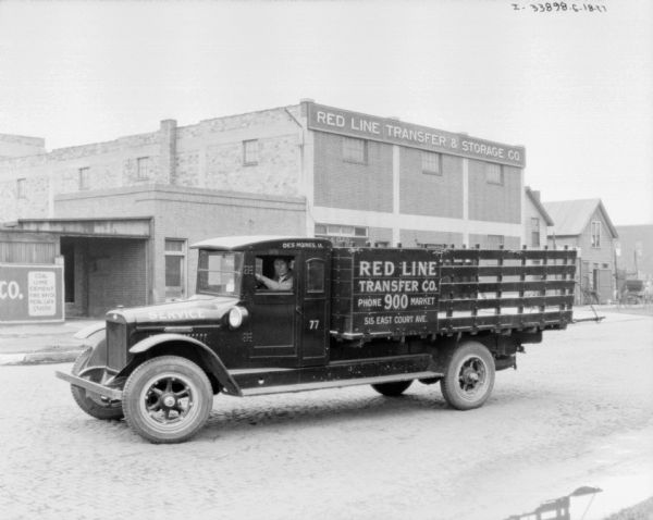 View across street towards a man sitting in the driver's seat of a delivery truck for Red Line Transfer Co. In the background is the Red Line Transfer & Storage Co. building.