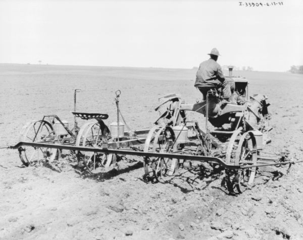 Three-quarter view from right rear of a man driving a Farmall tractor pulling a planter in a field.