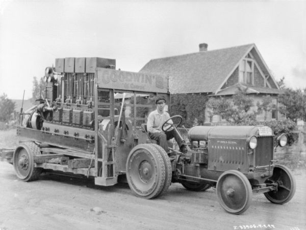 Three-quarter view from front right of a man sitting on a McCormick-Deering industrial tractor. The tractor is pulling a trailer full of equipment, and a sign on the front of the tractor reads: "Goodwin's."