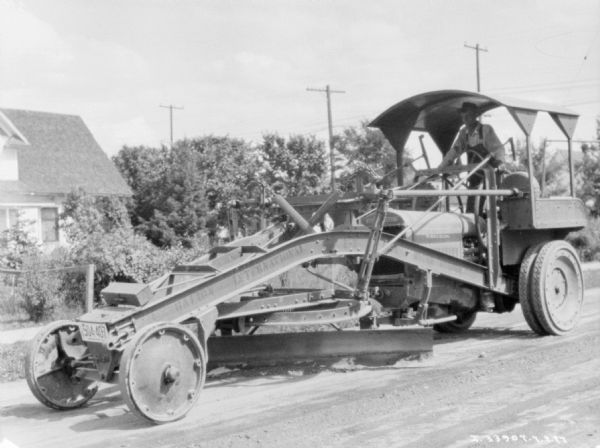 A man is driving a McCormick-Deering Tractor with a grader attached to the front to work on a road.