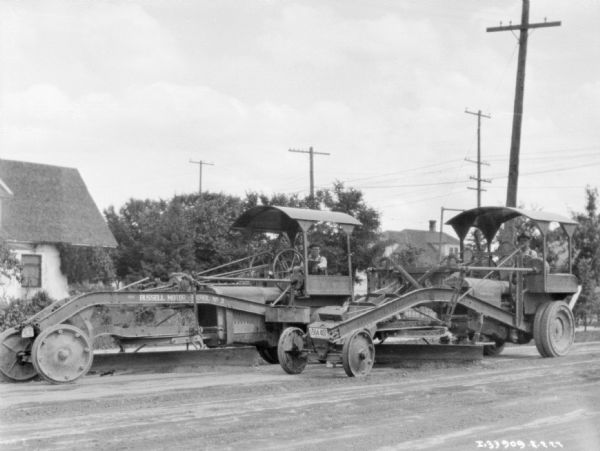 Two men with two road graders on a road. The grader on the left has a painted sign that reads: "Russel Motor," and the one on the right reads "_ International."