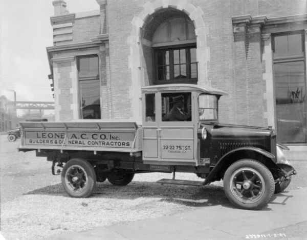 View of right side of delivery truck, with a painted sign along the truck bed that reads: "Leone A.C. Co. Inc, Builders & General Contractors. A man is is sitting in the driver's seat, and a large brick building with an arched entrance is just behind the truck.