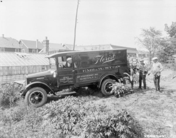 A man is sitting in the driver's seat of a floral delivery truck. The driver is looking back towards men loading plants into the back of the truck. The sign painted on the side of the truck reads: "Florist, Emil A. Stein." There is a greenhouse is the background on the left.