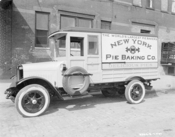 Driver's side view of delivery truck parked along a curb near a building. The sign painted on the side of the truck reads: "The World's Largest Pie Bakers, New York Pie Baking Co., Delicious Pies." A spare tire is mounted in the side of the truck on the running board under the driver's side window.