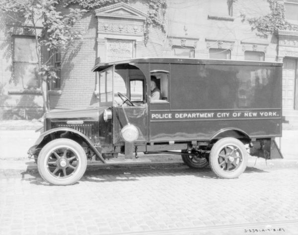 Driver's side view of a police department truck parked on a street. A man is sitting in the driver's seat, and the sign on the side of the truck reads: "Police Department City of New York." A large brick building behind the truck has a sign that reads, in part: "New York Architectural _."