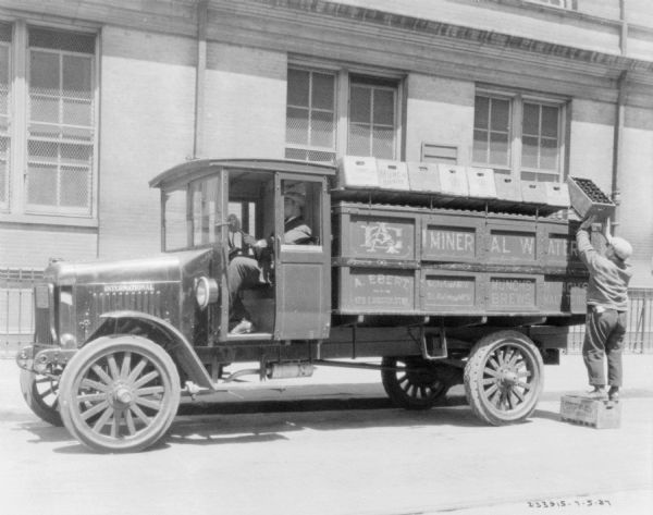 View across street towards a man sitting in the driver's seat of a beverage delivery truck parked along a curb in front of a large brick building. On the right a man standing on a wooden crate is loading or unloading a wooden crate of bottles at the side of the truck. A sign on the side of the truck reads: "A. Ebert."