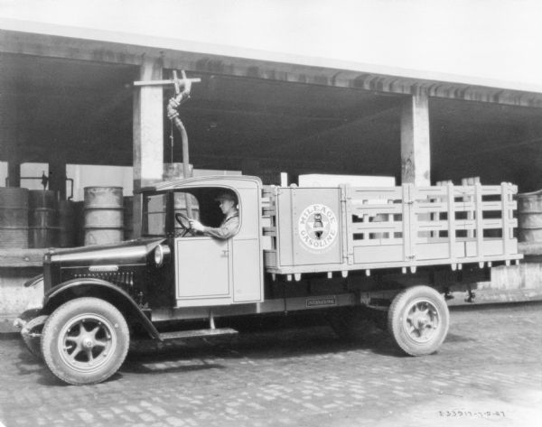 Driver's side view of a gasoline delivery truck. A man is sitting in the driver's seat. The logo on the side of the truck reads: "Mileage Gasoline." The truck is parked alongside what may be a loading dock, with barrels stored under the open sided roof.