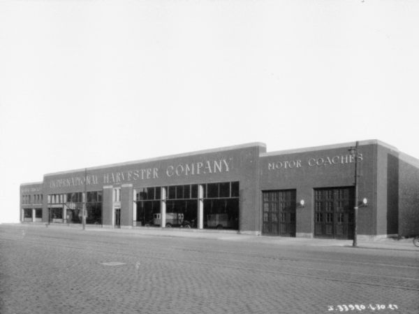 View across street towards an International Harvester Company dealership. Vehicles are on display in a showroom inside the dealership behind large show windows. On the right are garage doors with a sign for Motor Coaches above.