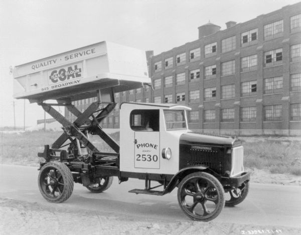 Right side view of a coal delivery truck parked outdoors. The coal box is raised high above the bed and cab roof `of the truck with a scissor lift. The sign on the coal box reads: "Quality • Service, Isaac Bloom Coal."
