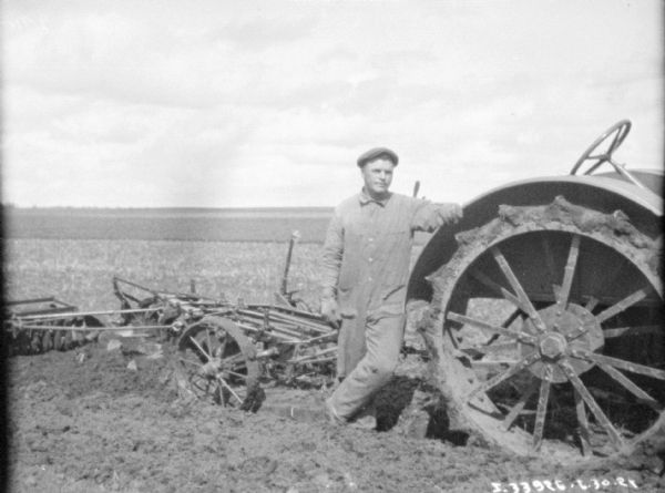 A man is standing near the right rear wheel of a tractor. The tractor is hitched to a plow and soil pulverizer.