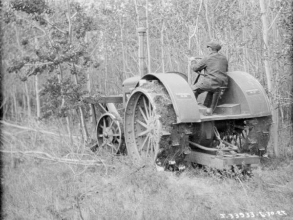 Three-quarter view from left rear of a man driving a McCormick-Deering tractor with a wood frame attached to the front for knocking down trees.