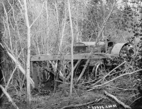 View through trees towards a man driving a Titan 15-30 McCormick-Deering tractor for knocking down trees with a wood frame attached to the front for knocking down trees.