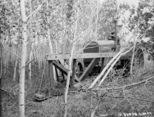 View through trees towards a man driving a Titan 15-30 McCormick-Deering tractor for knocking down trees with a wood frame attached to the front.