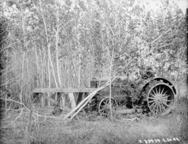 View through trees towards a man driving a Titan 15-30 McCormick-Deering tractor for knocking down trees with a wood frame attached to the front.