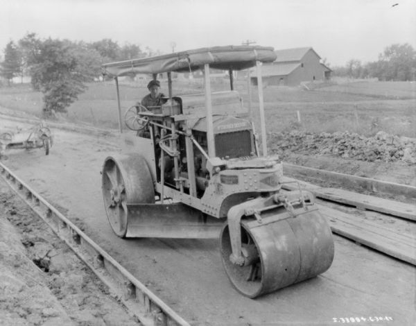 Slightly elevated view towards the front of a black top flattener. A man is driving the McCormick-Deering machine, and in the background are farm buildings.