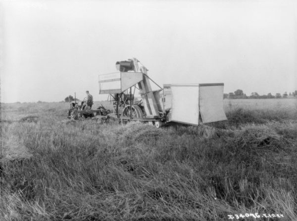Three-quarter view from rear of a man driving a Farmall tractor to pull a harvester-thresher in a field.