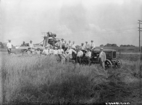 View of a group of men and children posing, on and around a tractor pulling a harvester thresher. Many of the men are wearing white shirts and neckties, 