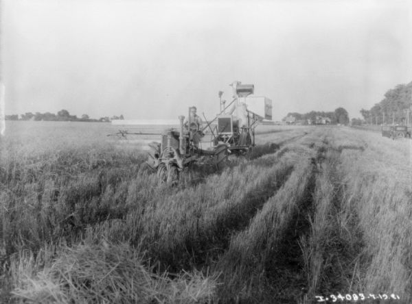 Three-quarter view from front of a man driving a Farmall tractor to pull a harvester-thresher in a field. There is an automobile in the field on the right, and farm buildings are in the distance.