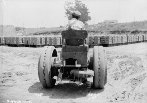 Rear view of a man driving an industrial tractor in a warehouse yard. Pallets of bricks are in the background.