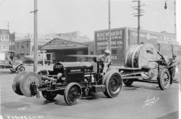 Man driving a tractor. There is a wood spool on the front of the tractor, and a larger wood spool on a trailer on the back.