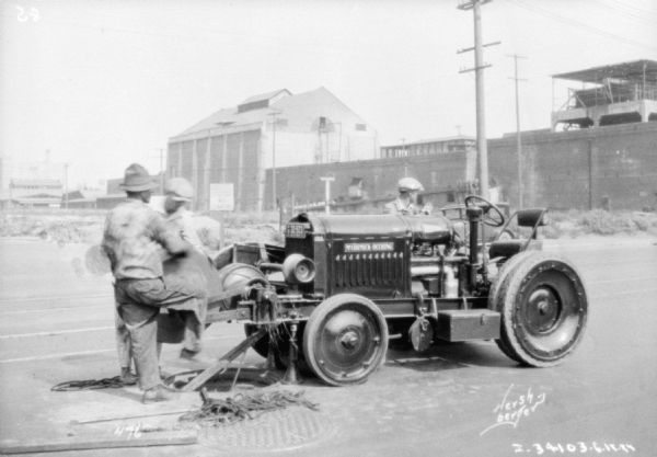 Two men are working with machinery on the front of an industrial McCormick-Deering tractor. Another man is standing behind the tractor.