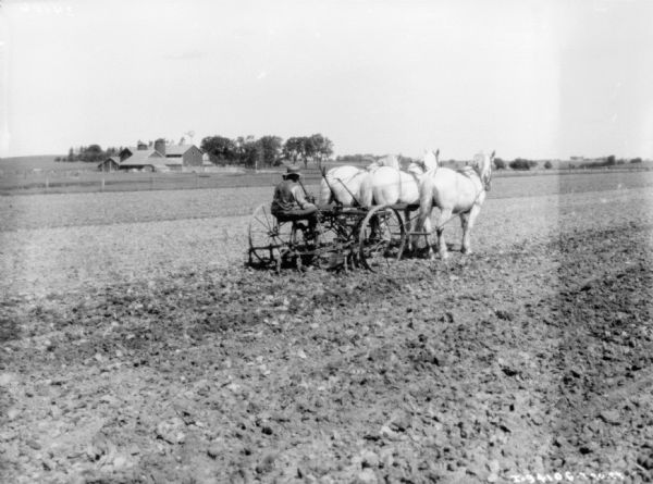 View across field towards a man riding on a horse-drawn cultivator. There is a farm with silo's and a windmill among trees in the background.