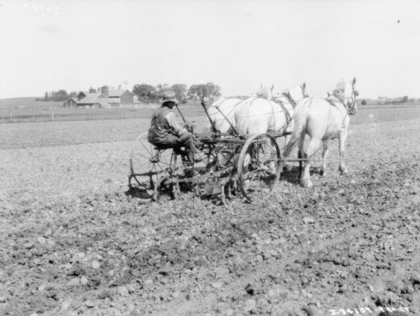 View across field towards a man riding on a horse-drawn cultivator. There is a farm with silo's and a windmill among trees in the background.