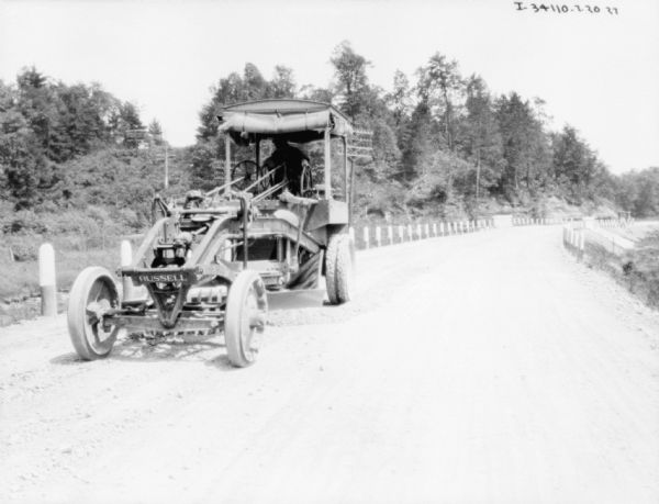 Front view of a grader. A man is operating the grader on a road with painted wooden guard rails. The sign on the front of the grader reads: "Russell."