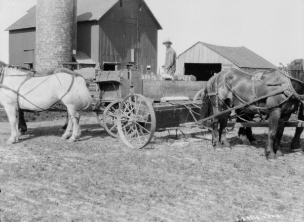 Man standing in horse-drawn wagon loading a horse-drawn manure spreader. There is a silo and barn in the background.