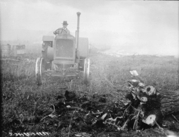 A man is driving a tractor in a field. There are tree stumps in the foreground.
