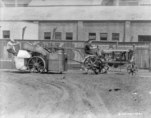 View across yard towards a man driving a Farmall tractor that is pulling another man sitting on the back of a cotton picker. In the background is a fence, and behind the fence are brick factory buildings.