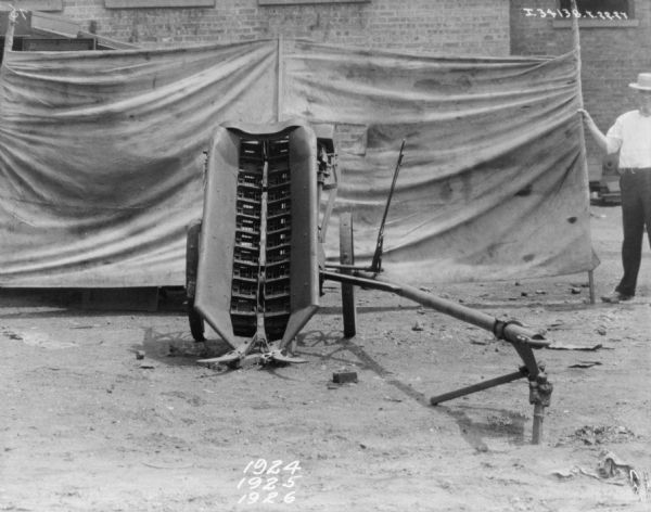Cotton picker set up outdoors. A man is standing in the background on the right holding up a sheet as a backdrop. There is a brick building in the background.
