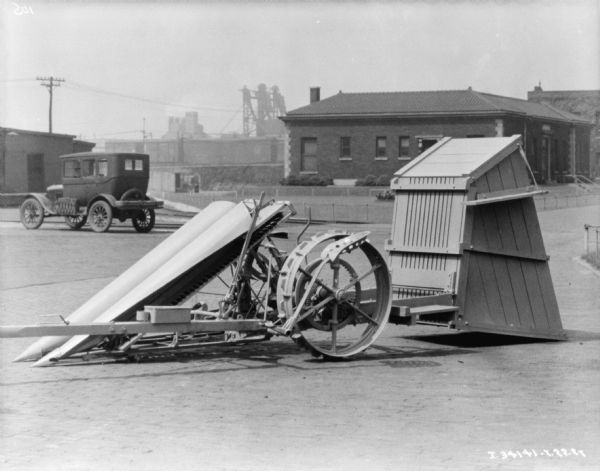 A cotton picker is parked outdoors with attached carrier. An automobile is parked in the background. Buildings, some industrial looking, are in the far background.