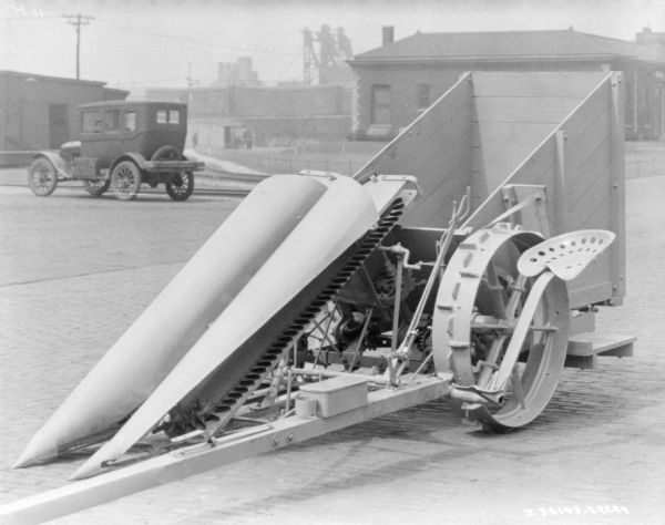 Cotton picker parked outdoors. An automobile is parked in the background. Buildings, some industrial looking, are in the far background.