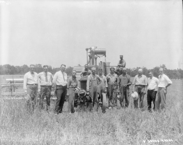 A large group of men are posing in front of a man sitting on a tractor pulling a harvester thresher in a field.
